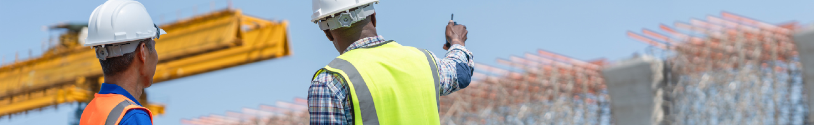 Two OT engineers in safety vests and hard hats reviewing critical infrastructure at an active construction site with industrial cranes and structural framework.